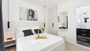 Un dormitorio moderno con una cama grande, una mesita de noche con un libro, una botella de vino y una bandeja de uvas. Hay un cuadro en la pared y un espejo sobre la cama.