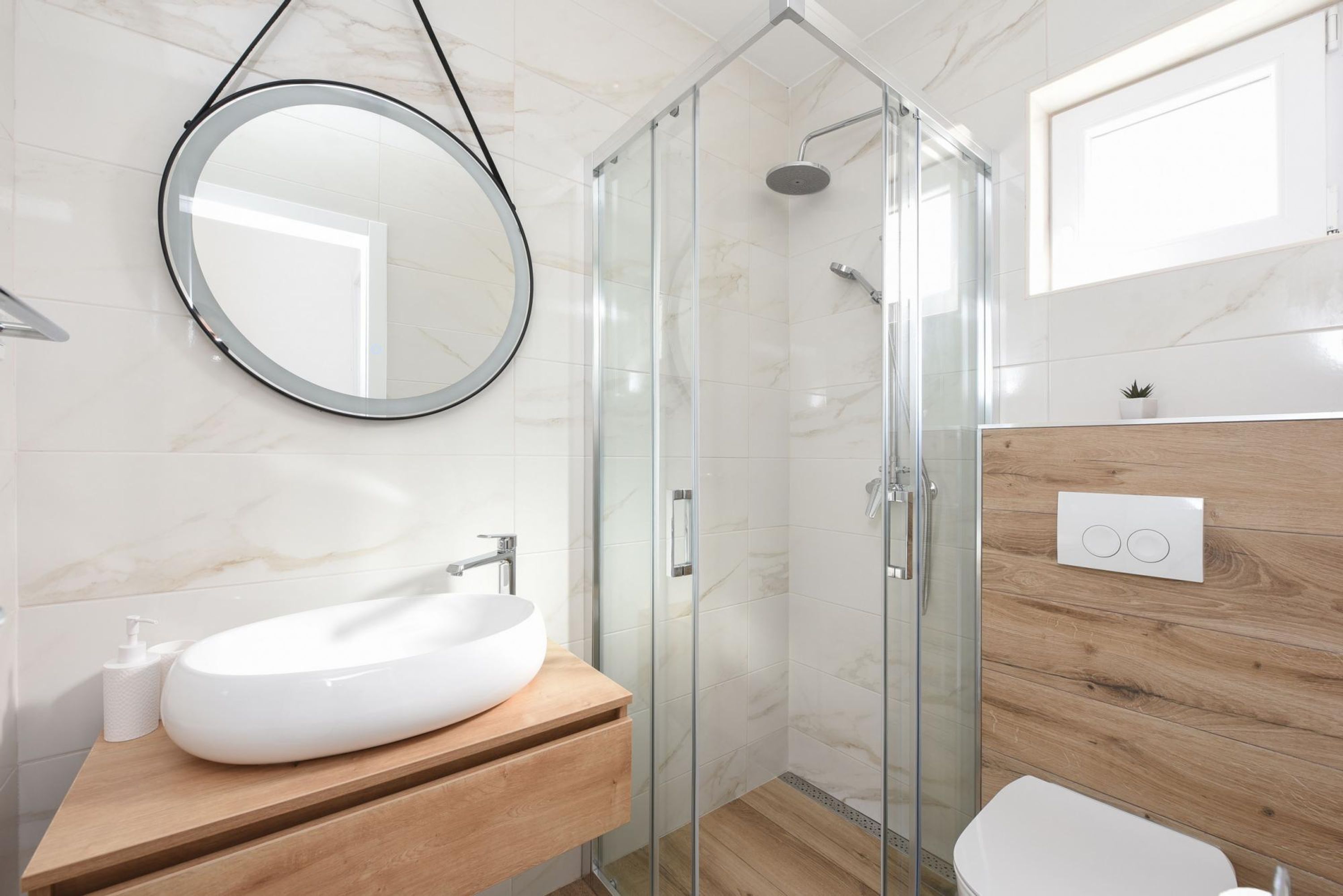 Ein modernes Badezimmer mit einem weißen Waschbecken, einem Holz-Waschtisch, einer Glasdusche und einem runden Spiegel.
