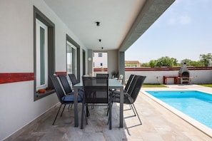 An outdoor patio with a pool, a table with chairs, and a barbecue grill.