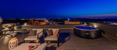 A rooftop terrace with outdoor seating, a hot tub, and a view of the city at dusk.