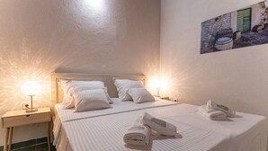 Una habitación de hotel con dos camas, cada una con ropa blanca y almohadas, una mesita de noche con lámpara, y un cuadro en la pared.
