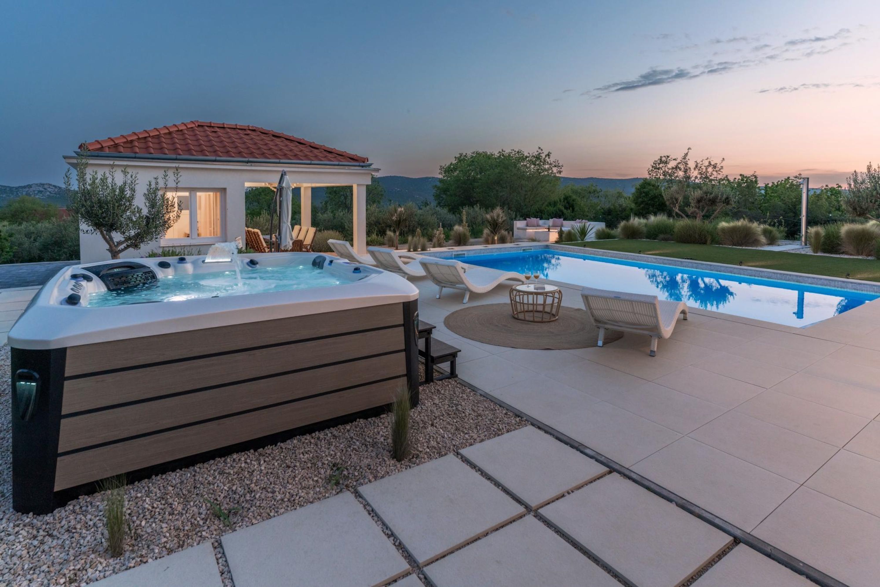 Un jacuzzi y una piscina con sillas de descanso, rodeados por un jardín ajardinado y un pabellón.