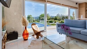 Un salon moderne avec une table basse en verre, un canapé bleu et une vue sur une piscine et un espace verdoyant.