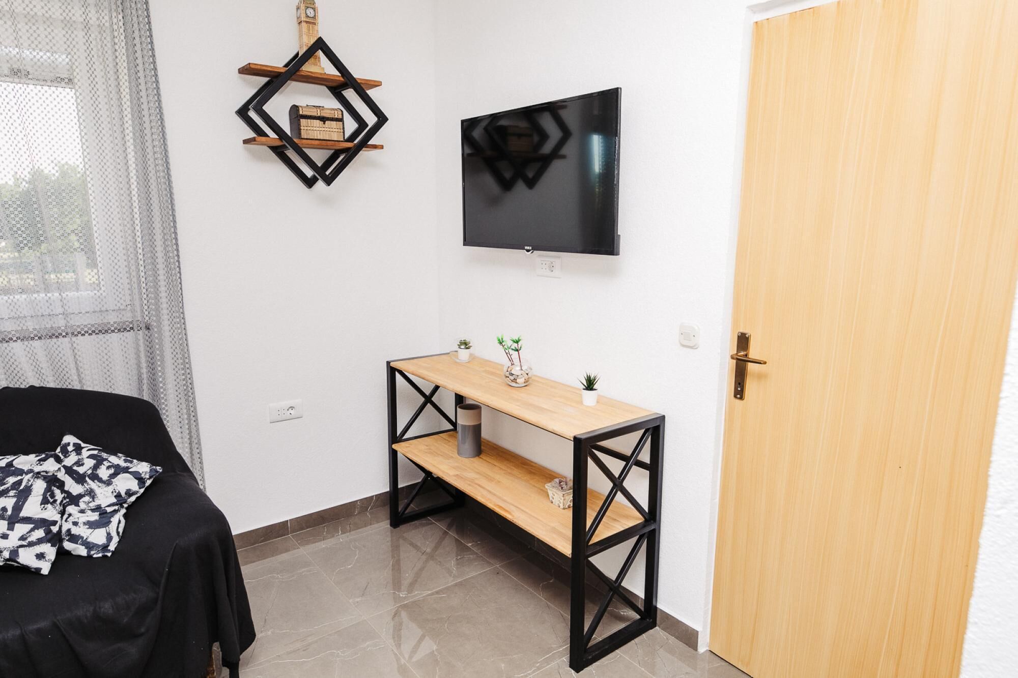 Ein kleines Zimmer mit einem Bett, einem Holztisch mit Regalen, einem Fernseher und einer Tür.
