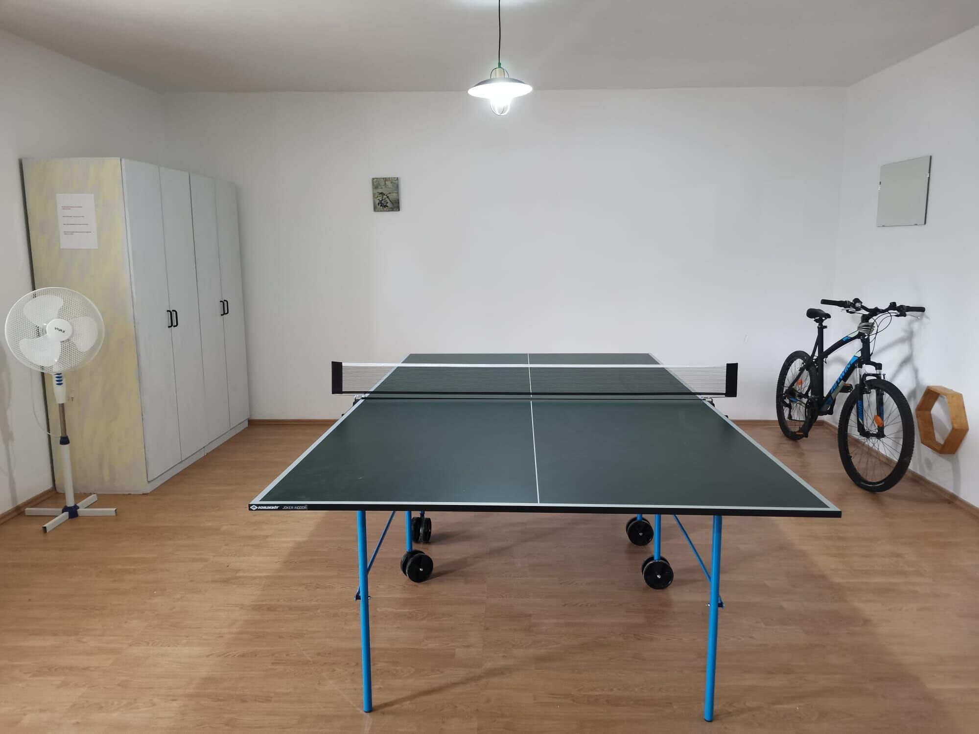 Una sala con pavimento in legno, un ventilatore a piantana e una bicicletta, con un tavolo da ping pong.