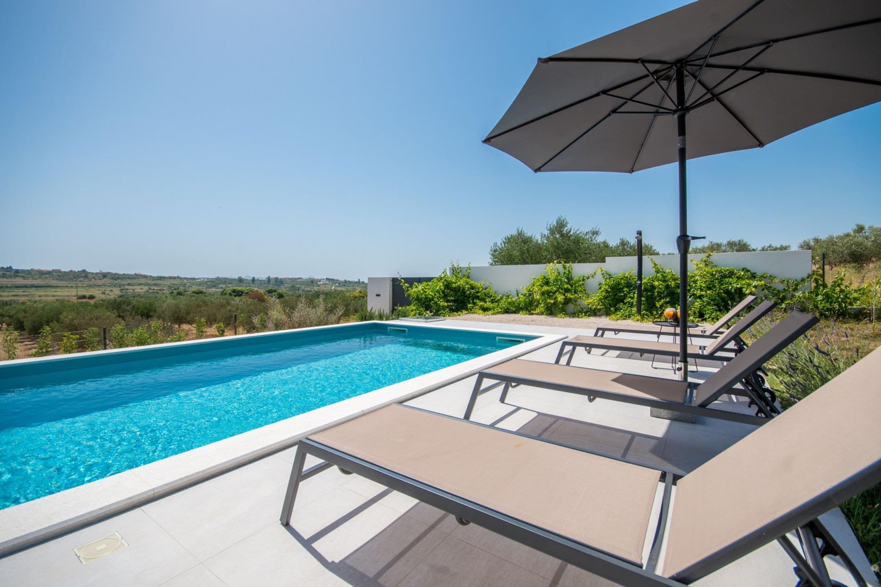 Une piscine entourée de verdure, avec des chaises longues et un parasol, sous un ciel dégagé.