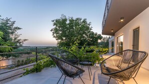 A balcony with two black wire-framed chairs and a small table, overlooking a lush garden and distant landscape.