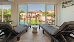 Un balcon avec des fauteuils de jardin en osier, une petite table avec une bouteille et un verre, et une vue sur une piscine et des maisons au loin.