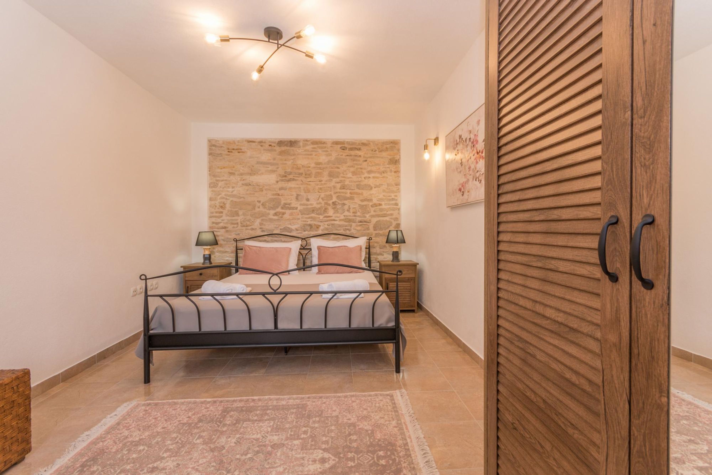 A bedroom with a bed, bedside tables, a wooden wardrobe, a stone wall, and a ceiling light fixture.