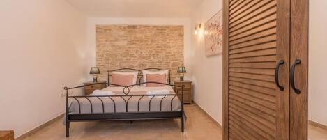 A bedroom with a bed, bedside tables, a wooden wardrobe, a stone wall, and a ceiling light fixture.