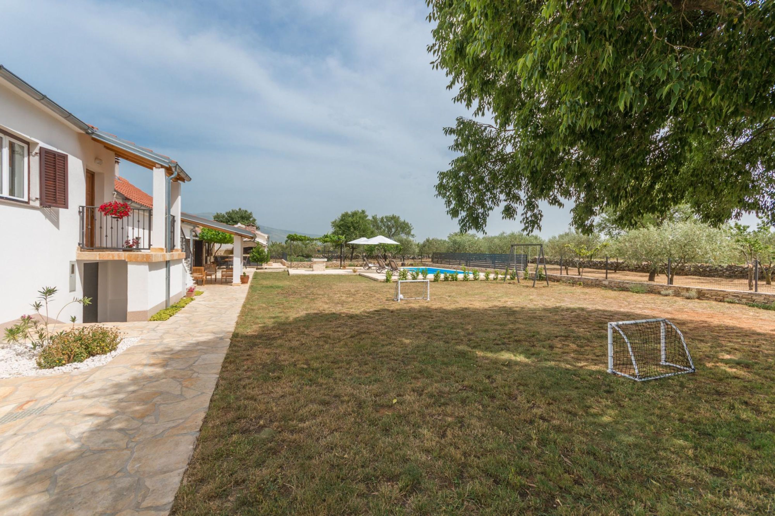 A residential property with a stone pathway leading to a white house with a balcony, a swimming pool, and a soccer goal in the yard.