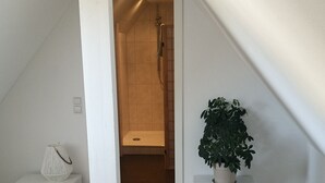 A modern bedroom with a sloped ceiling, a wooden floor, a white bedside table with drawers, a potted plant, and a shower area visible through an open door.