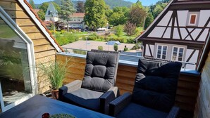 A balcony with two cushioned chairs and a table, overlooking a scenic view of a village and a lake.