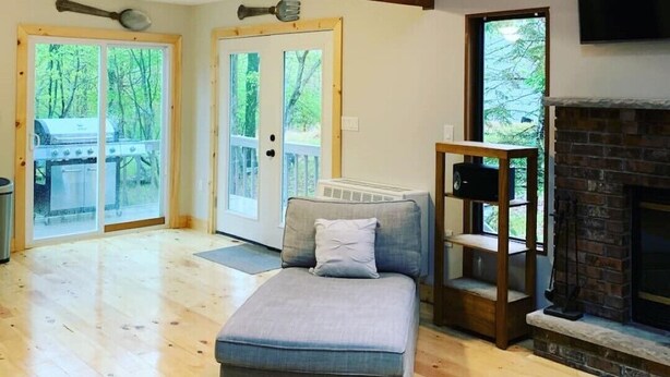 Uma sala de estar ampla, com teto alto, escada de madeira, lareira e uma grande janela com vista para um deck.