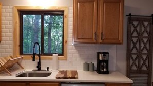 Uma cozinha com armários de madeira, lava-louças de aço inoxidável, cafeteira e janela com vista para árvores.