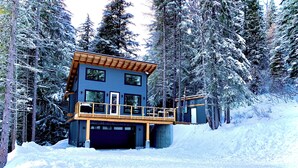 A two-story cabin with a wooden deck and a garage, surrounded by snow-covered trees.