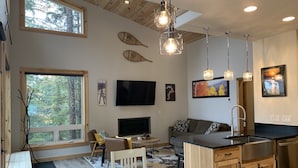 A modern kitchen with a large island, wooden cabinets, and a built-in oven. There is a dining area with a table and chairs, and a large window offering a view of the forest.