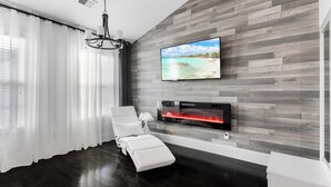 A modern living room with a large flat-screen TV mounted on a wooden wall, a fireplace with a red-lit surround, a white sofa, and a chandelier.