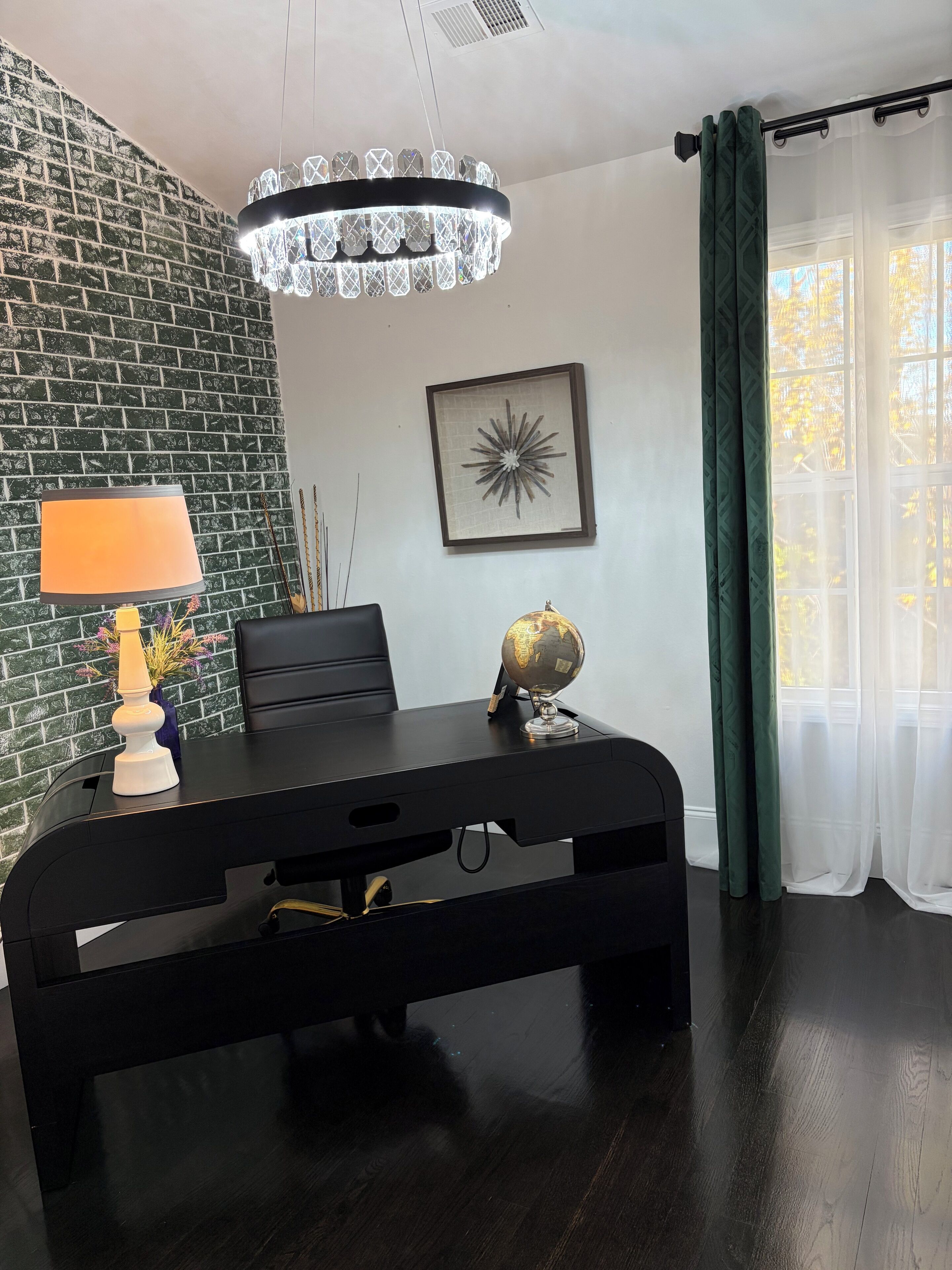 Una habitación con pared de ladrillo, un escritorio negro con un globo terráqueo y una obra de arte enmarcada, una lámpara y una araña.