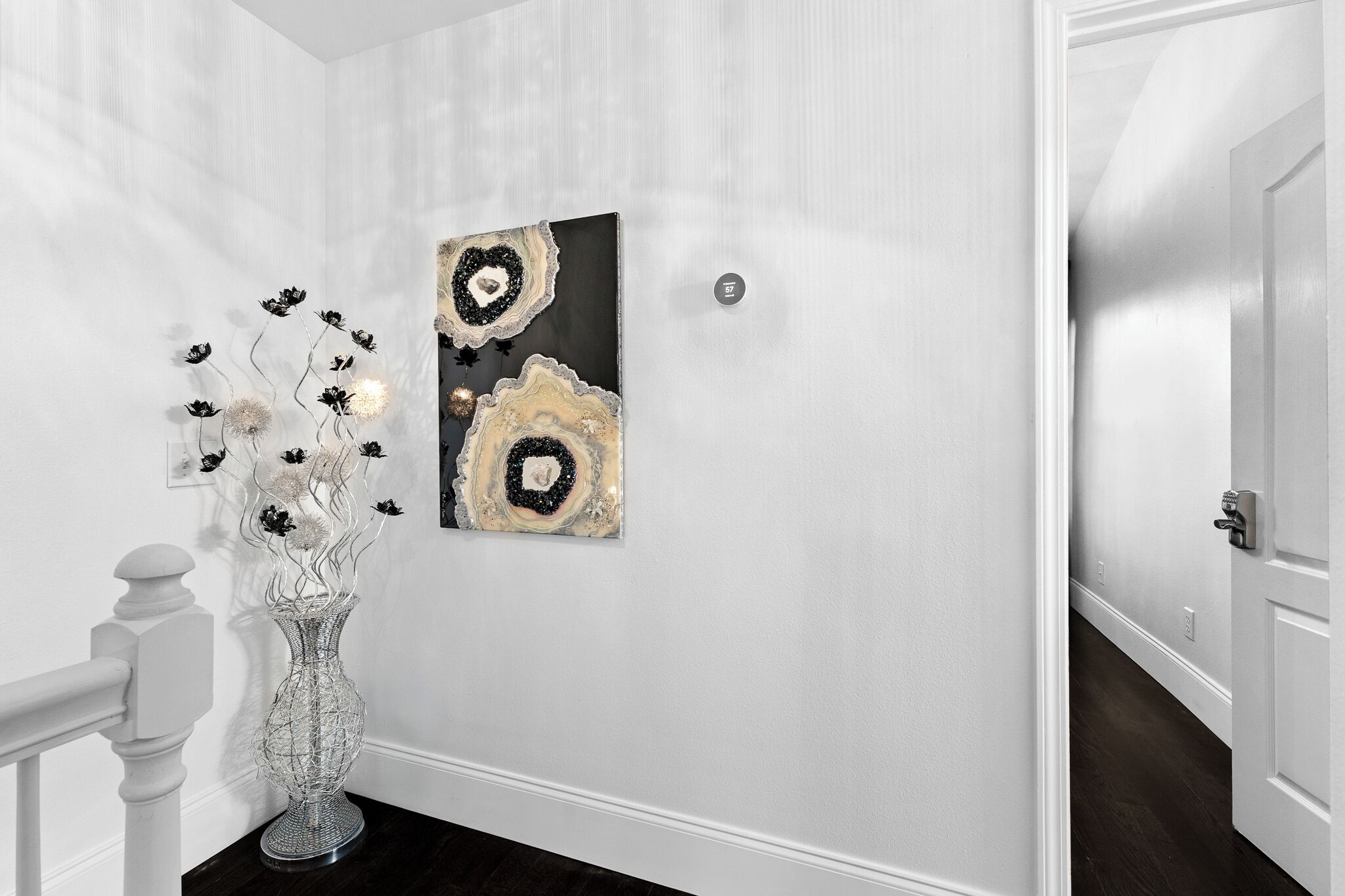 A hallway with a white wall, a black and gold abstract painting, and a clear glass vase with flowers.