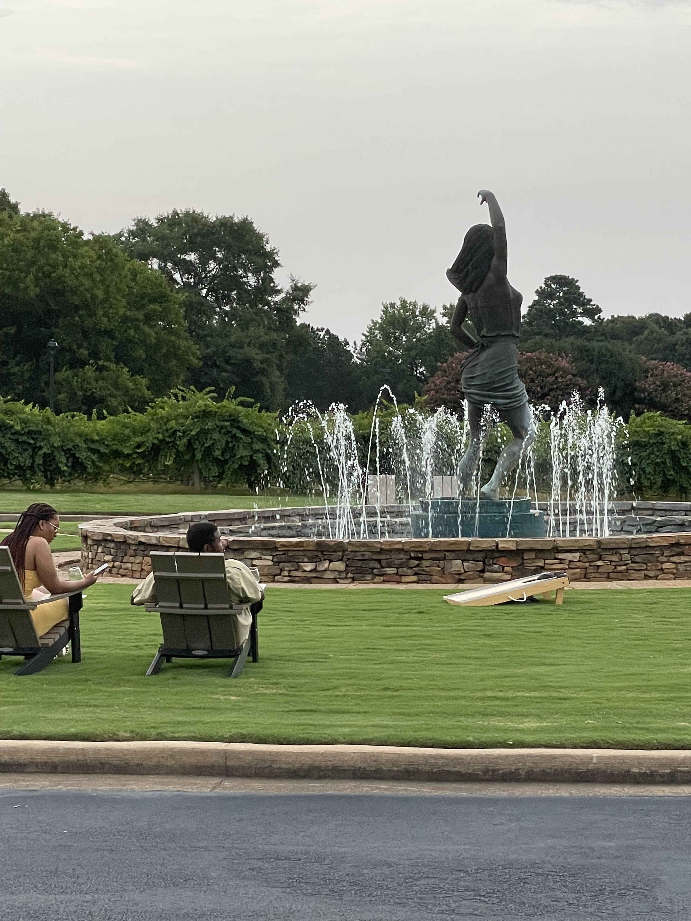 Eine Statue eines Wesens mit erhobener Arm in einem Park mit Brunnen, Bänken und einer Rasenfläche.