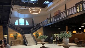 A spacious lobby with a high ceiling, skylights, and a central staircase. There are two large tables with potted plants, and a dining area with tables and chairs.