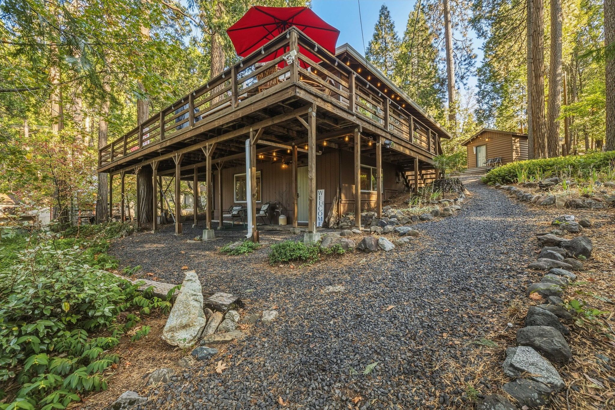 A wooden cabin with a porch, surrounded by trees and a gravel path.