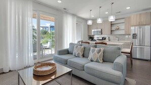 A modern living room with a grey sofa, a glass coffee table, and a kitchen area with a microwave, refrigerator, and wooden cabinets.