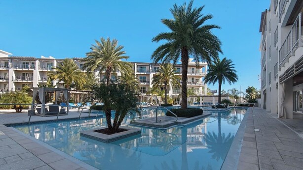 A swimming pool with palm trees, lounge chairs, and a building with balconies in the background.