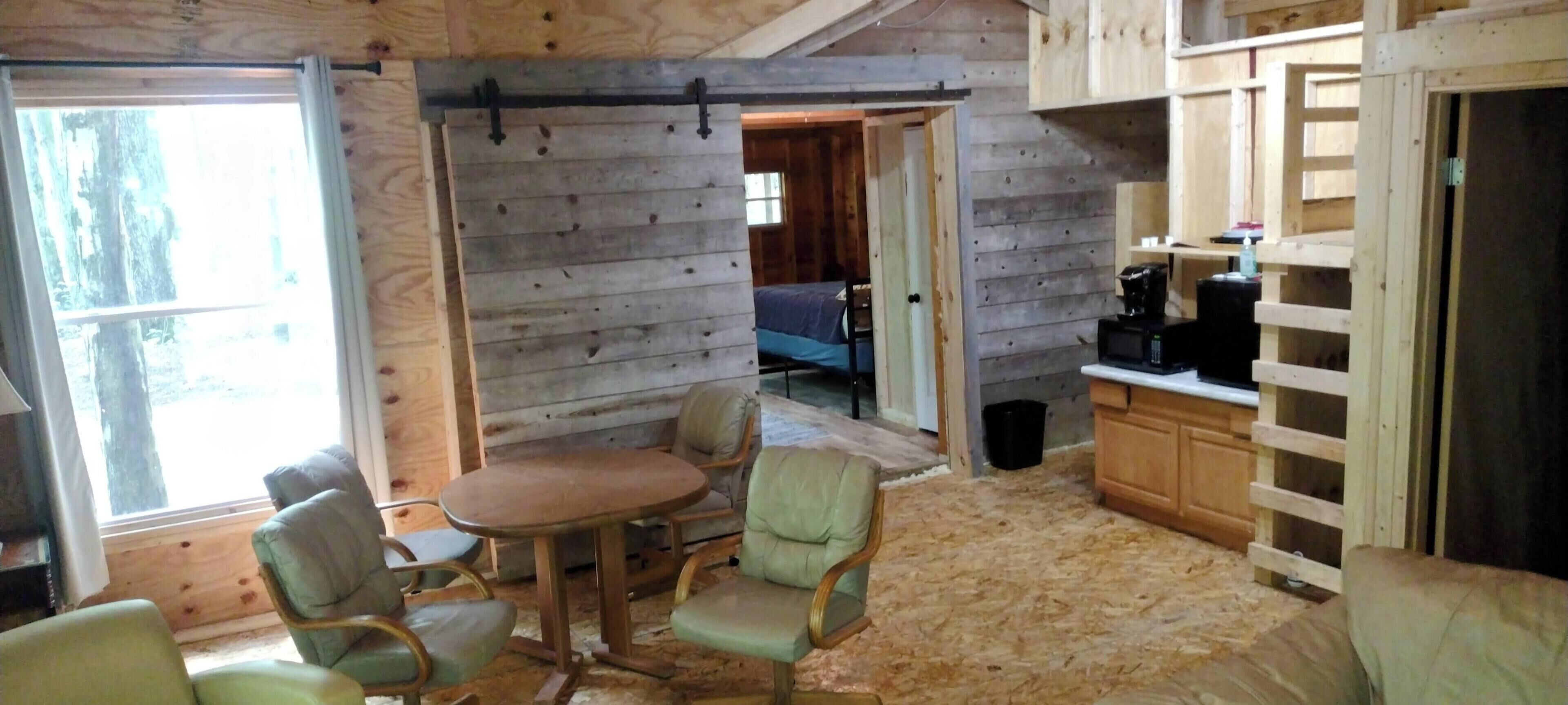 A cozy cabin interior with a wooden dining table and chairs, a kitchen area, and a view through an open door.