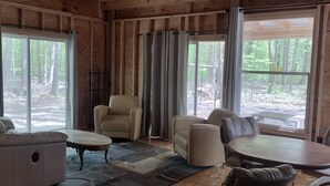 A log cabin interior with a sofa, armchairs, a small table, and large windows offering a view of a forested area.