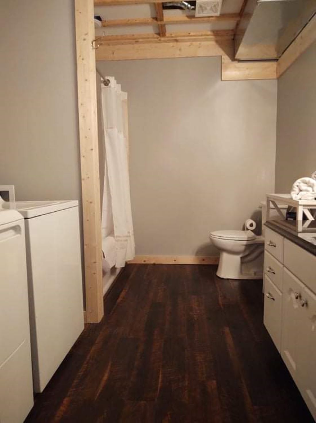 Een kleine, compacte badkamer met een houten plafond, een wit toilet, een douchegedeelte met een witte gordijn en een klein wastafeltje met spiegel.