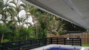 A covered swimming pool with lounge chairs, surrounded by a wooden fence and lush greenery.