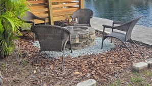 A wooden deck with wicker chairs and a stone fire pit by a pond.