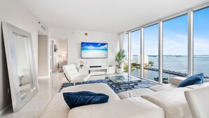 A modern living room with a large window, a flat-screen TV, and a view of the water. - ICON BRICKELL • CORNER TWO BEDROOM• OCEAN VIEW (Miami)