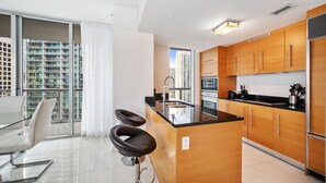 A modern kitchen with wooden cabinets, a black countertop, and a stainless steel refrigerator. There is a large window offering a view of high-rise buildings. - ICON BRICKELL • CORNER TWO BEDROOM• OCEAN VIEW (Miami)