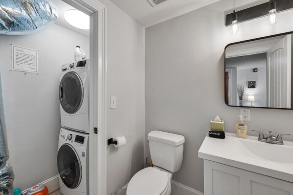 A compact bathroom with a washing machine, toilet, sink, and mirror.