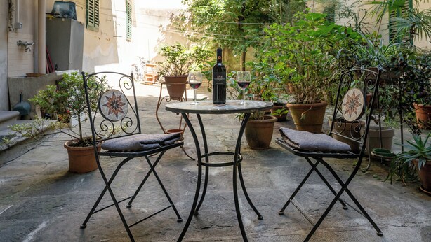 Eine Terrasse im Freien mit einem runden Tisch, zwei Stühlen, Topfpflanzen und einer Weinflasche.