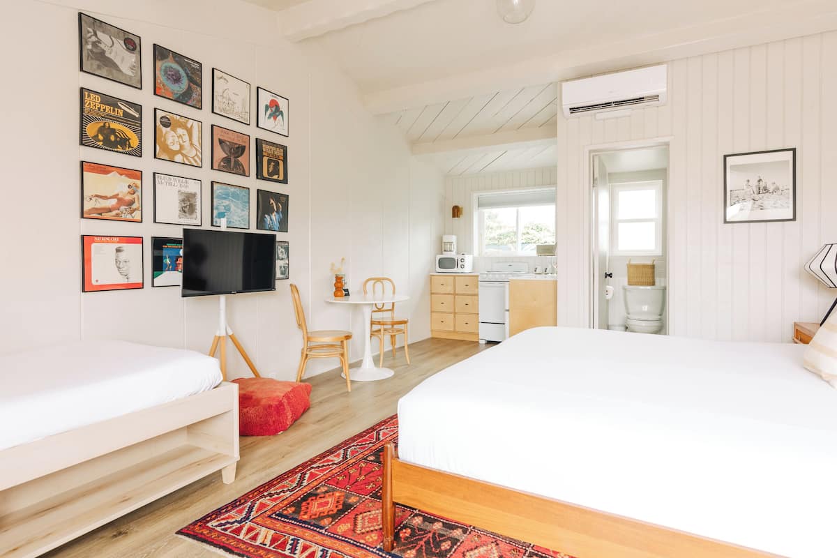 A bedroom with two beds, a TV, a small table with chairs, and a kitchen area visible through an open door.