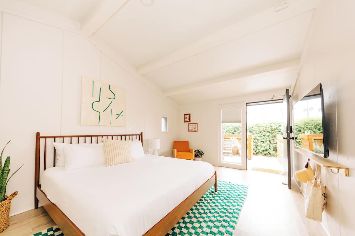 A bedroom with a large bed, a wooden headboard, a green and white checkered rug, a television mounted on the wall, and a sliding glass door leading to an outdoor area.