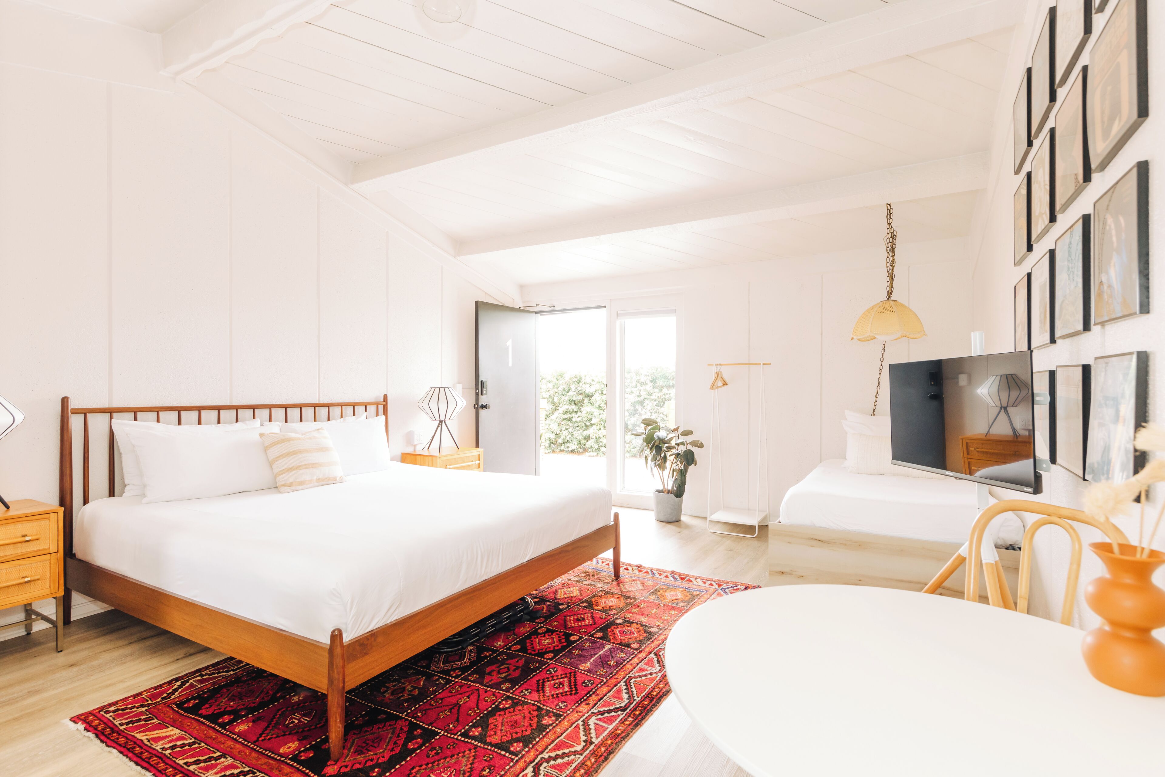 A modern bedroom with a wooden bed, a red patterned rug, a white bedside table, and a TV on a stand.