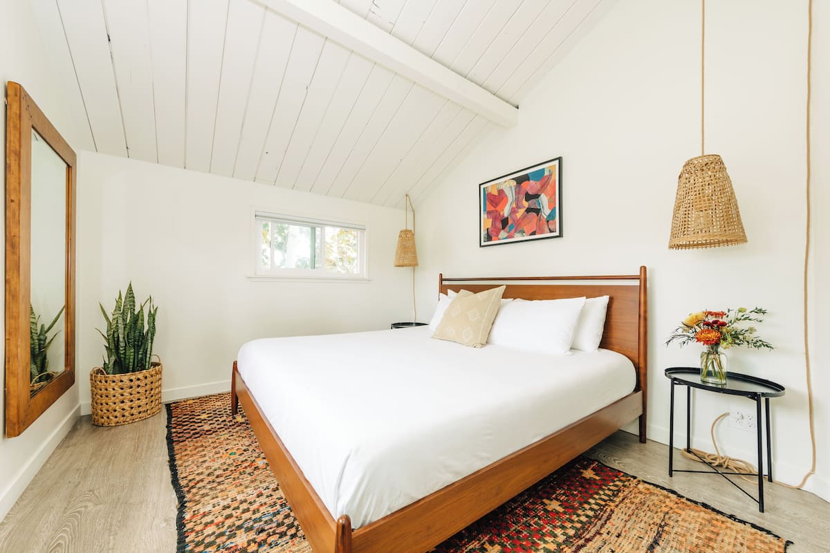 A bedroom with a wooden bed, a woven basket with plants, a colorful abstract painting, and a small table with a vase of flowers.