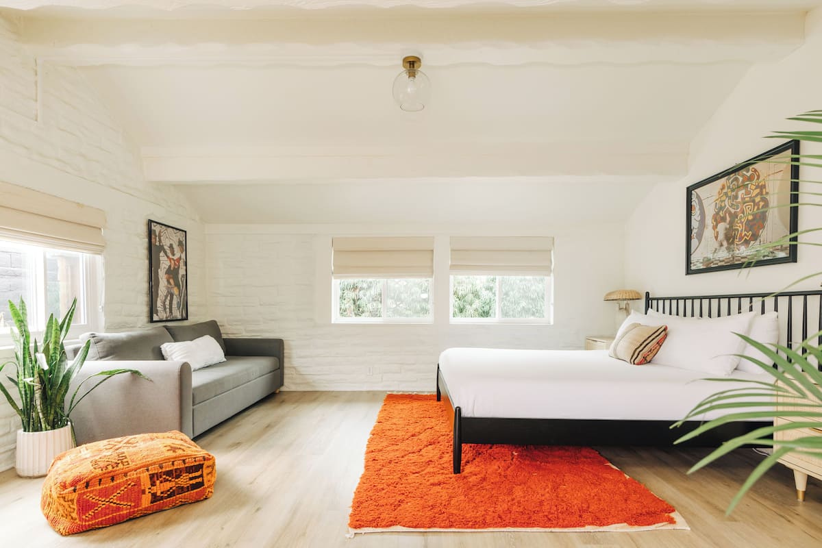A bedroom with a bed, a grey sofa, a red rug, and a potted plant.