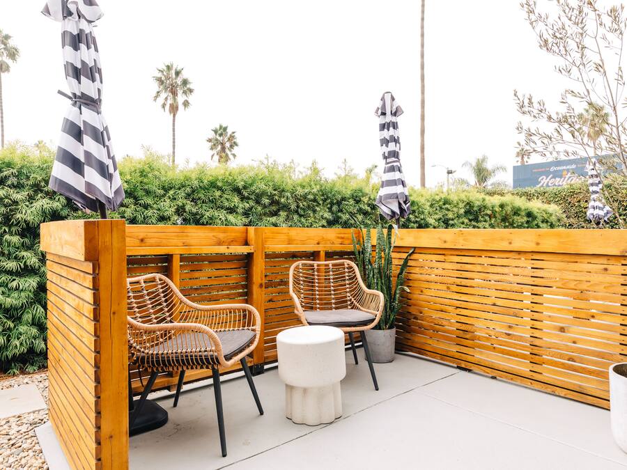 A wooden outdoor seating area with wicker chairs and a small round table, surrounded by a wooden fence and greenery.