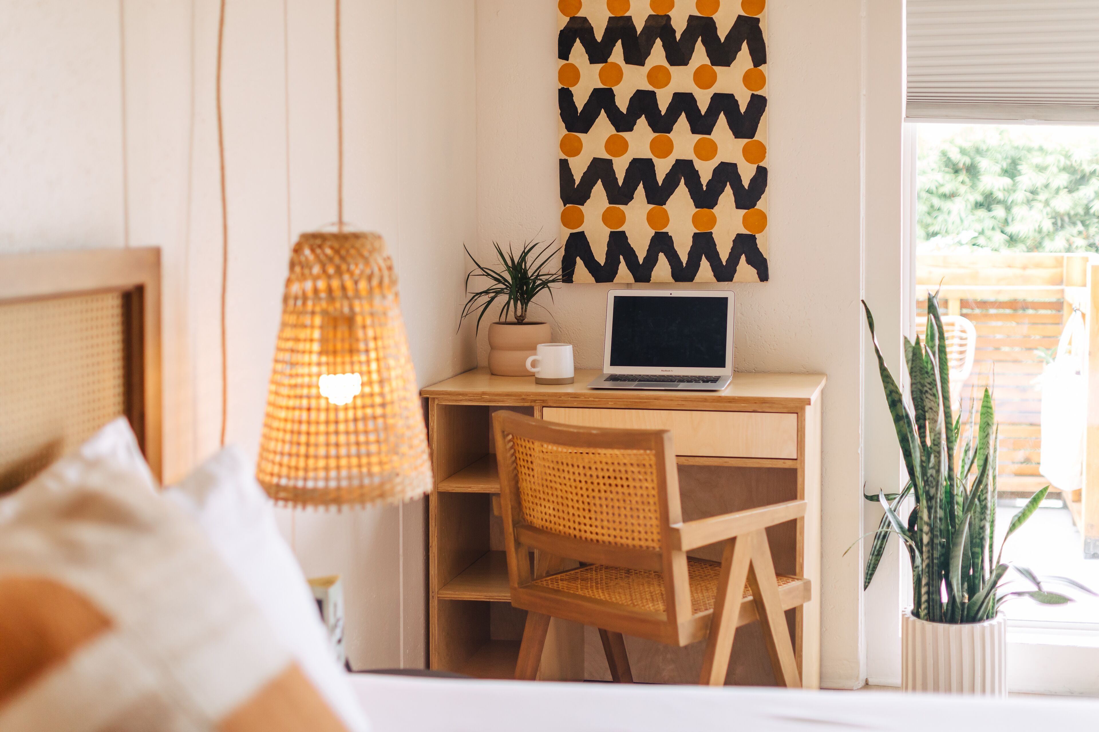 A cozy workspace with a laptop on a wooden desk, a woven chair, a hanging lamp, and a potted plant.