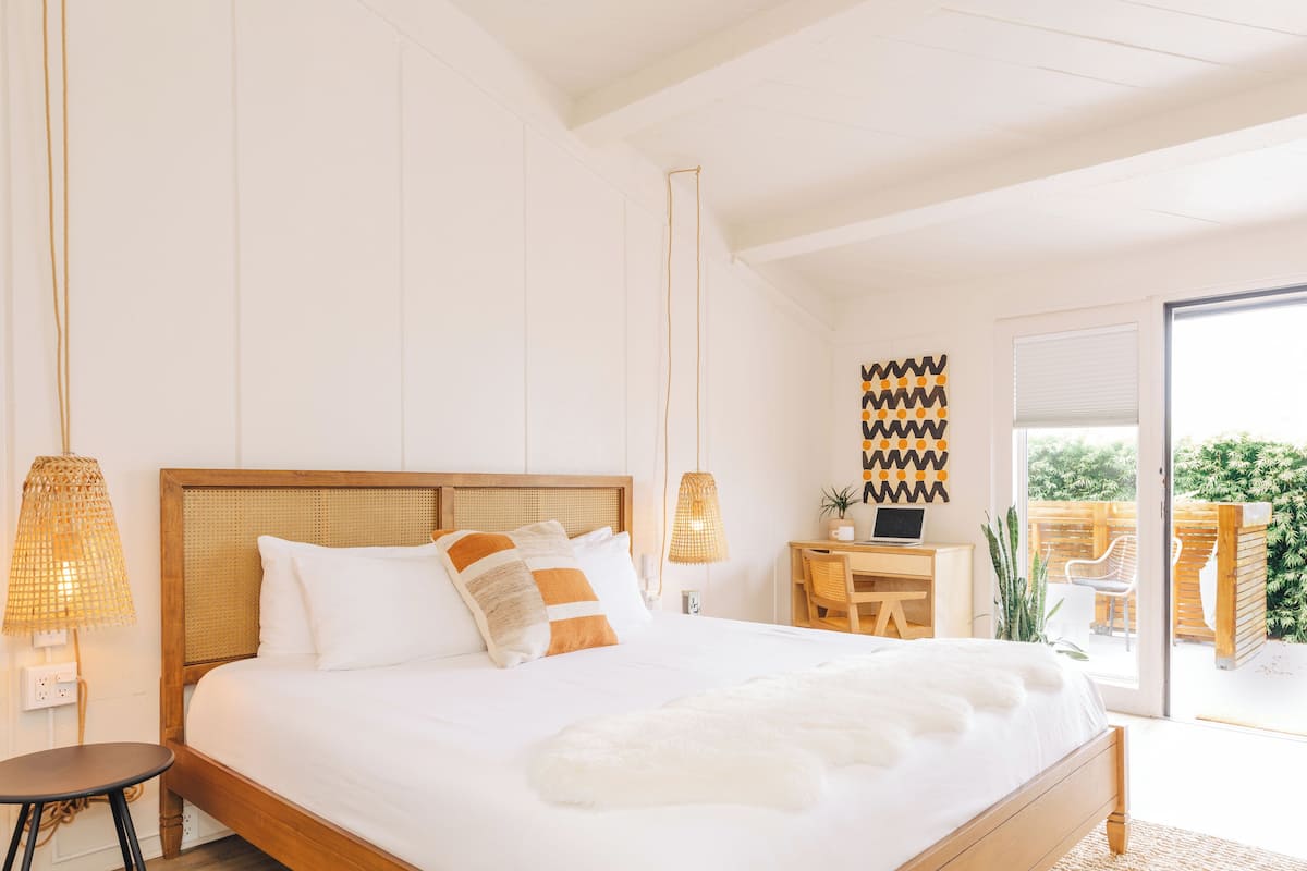 A bedroom with a large bed, a desk, and a chair. There is a window and a door leading to an outdoor area.