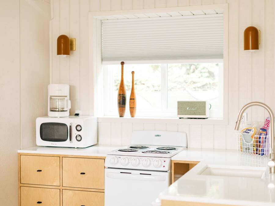 Una cocina con estufa blanca, microondas, armarios de madera y una ventana con persianas.