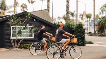 Two people riding bicycles on a street with a small building and palm trees in the background.