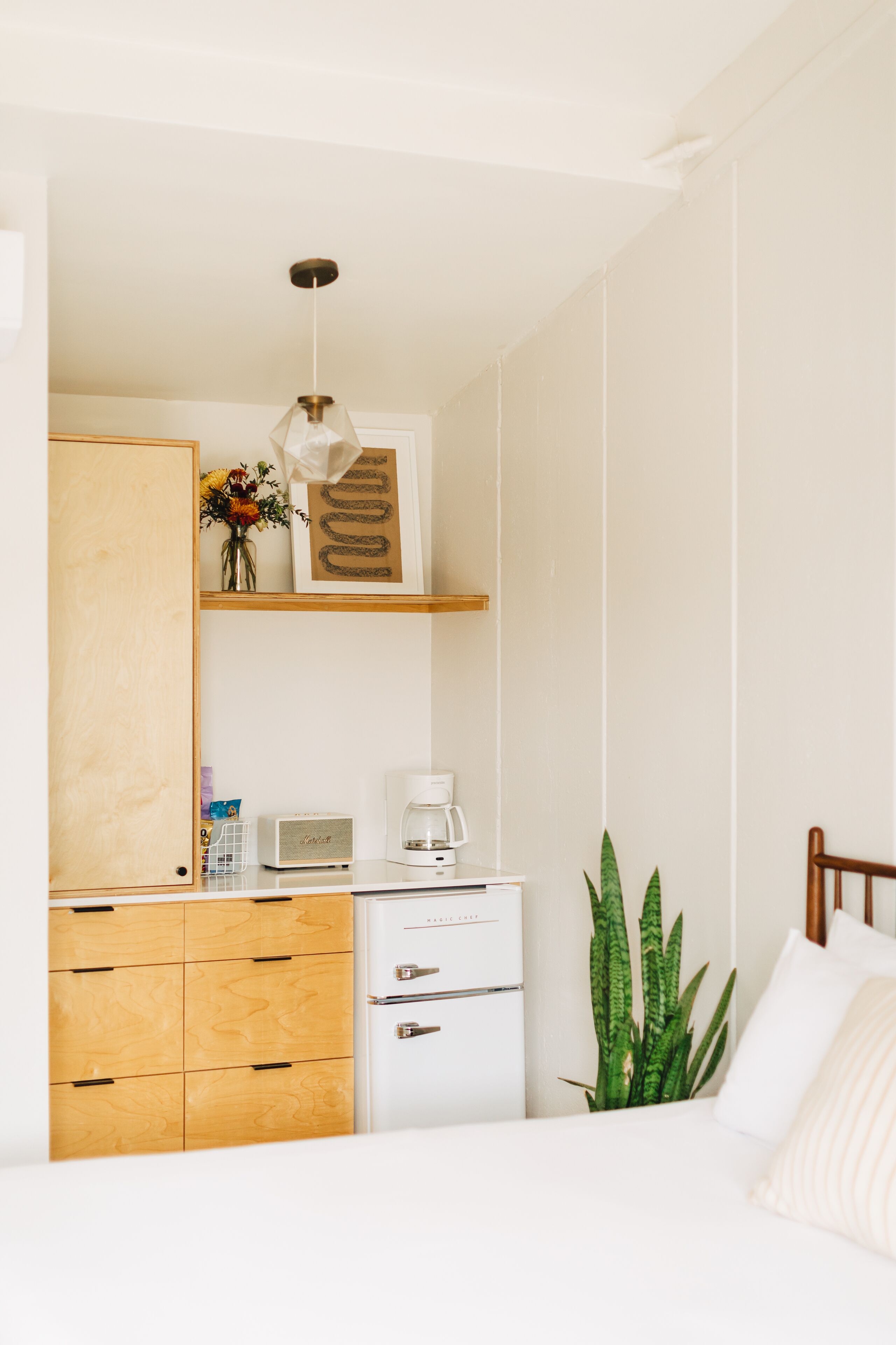 Un dormitorio con cama, armario de madera, escritorio con cafetera y una planta.