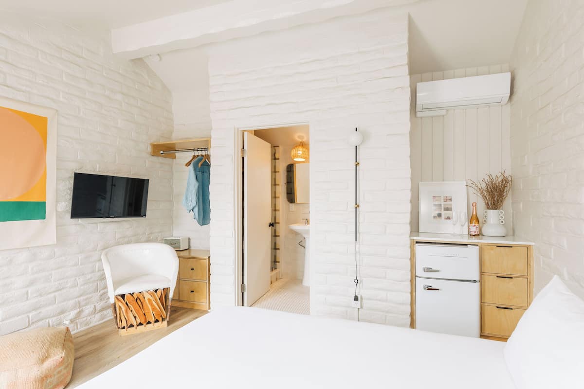 A modern bedroom with a white brick wall, a flat-screen TV, a bed, a wooden nightstand, a dresser, and a bathroom visible through an open door.